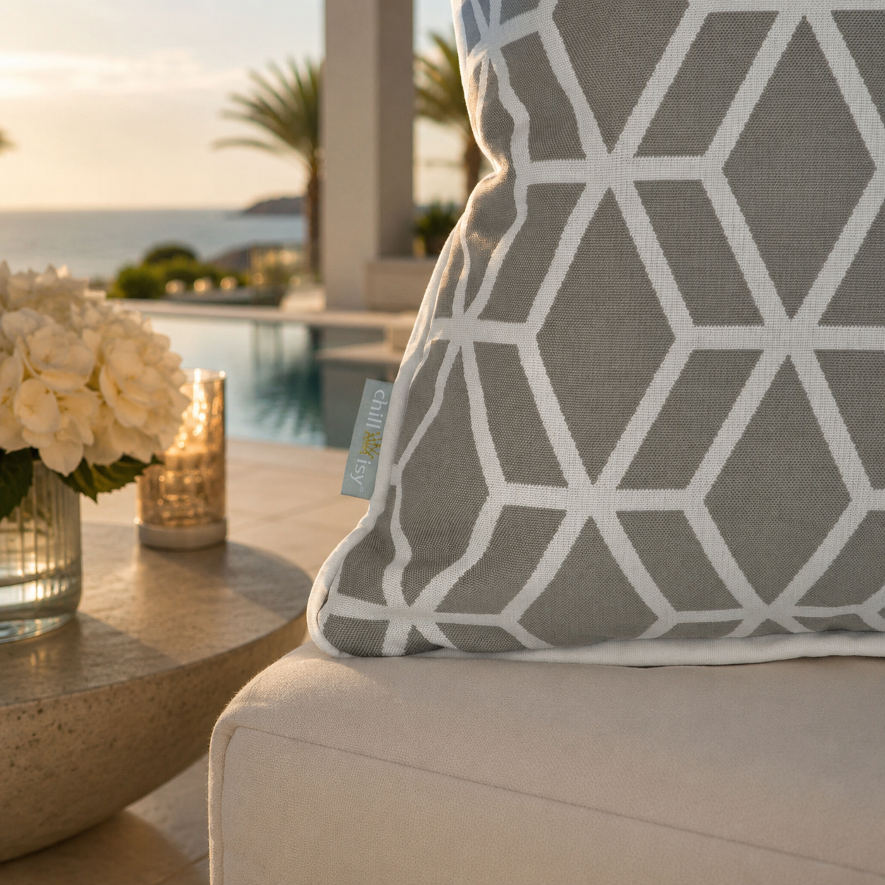 Detailaufnahme des chillisy® Designer Outdoor-Kissens Crystal Gold Hellgrau mit weißem geometrischem Muster, weißem Keder und Logo-Etikett auf einer luxuriösen Terrasse mit Pool und Meerblick – genäht in Deutschland