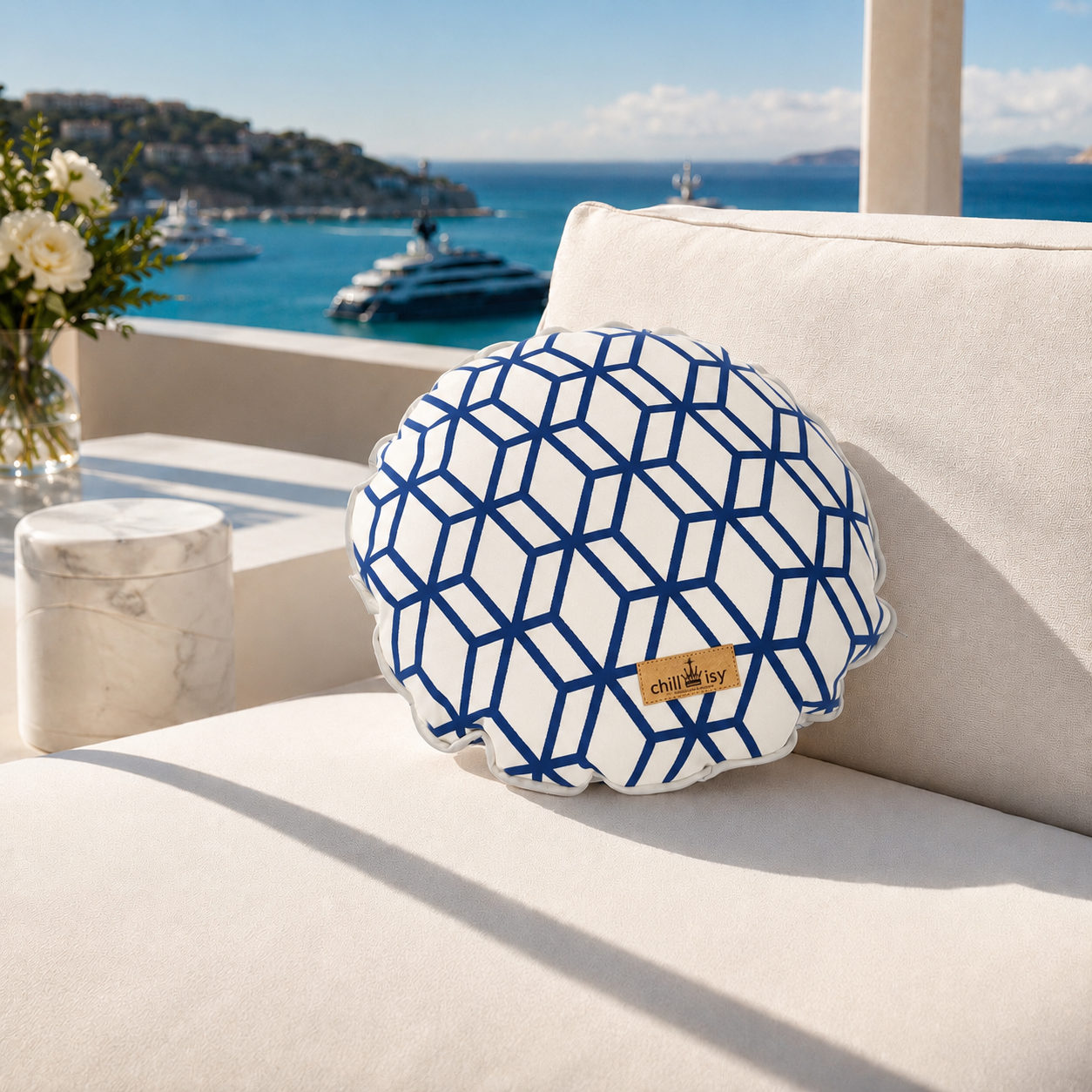 Designer-Rundkissen Crystal Gold von chillisy® in Weiß-Blau mit geometrischem Muster auf einer exklusiven Outdoor-Lounge mit Meerblick.