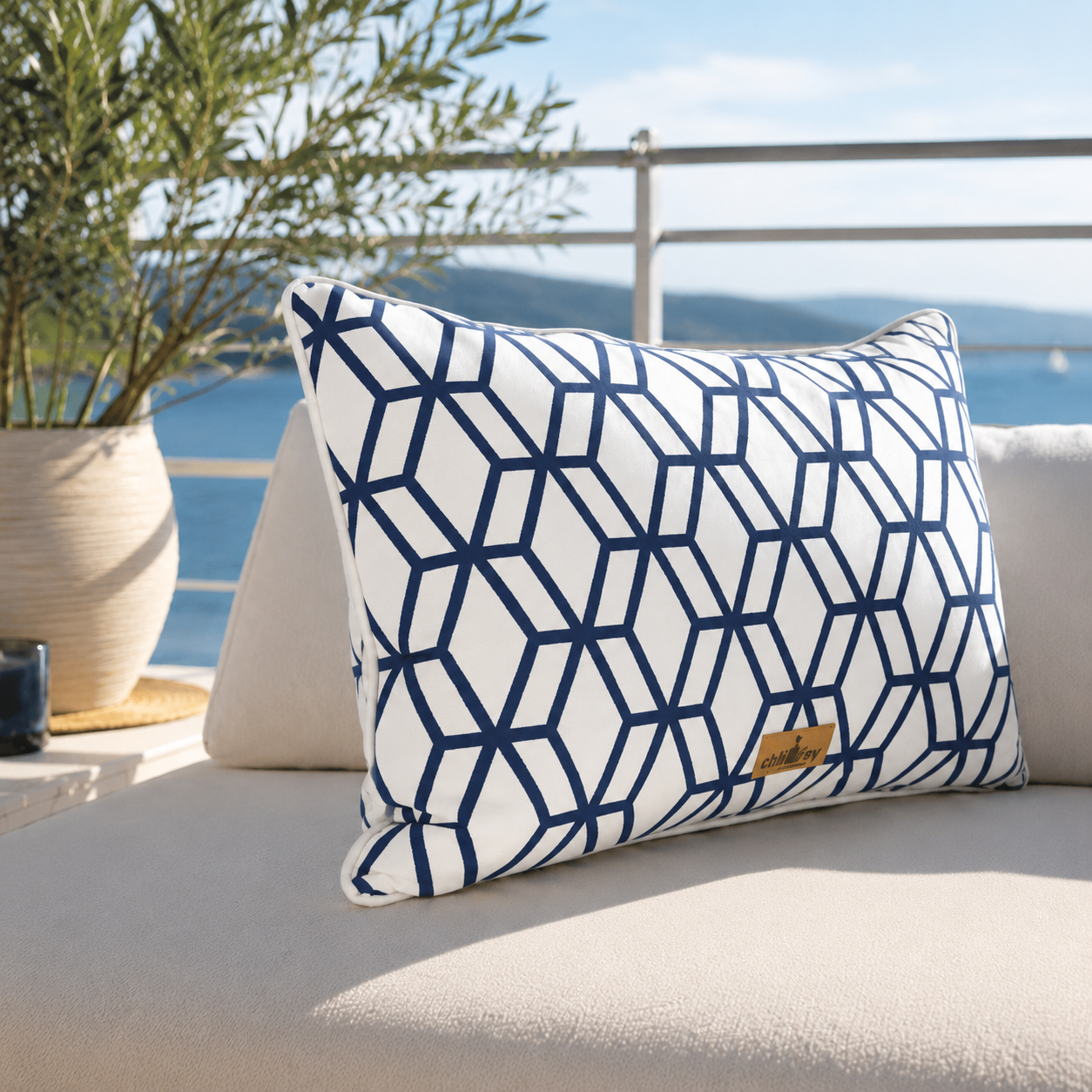 Luxuriöses Outdoor-Dekokissen von chillisy® mit weißem Stoff, marineblauem geometrischem Muster, weißem Keder und goldenem chillisy®-Label auf einer eleganten weißen Gartenlounge mit Meerblick. Wetterfestes Designerkissen für Garten, Terrasse, Pool und Yacht.

