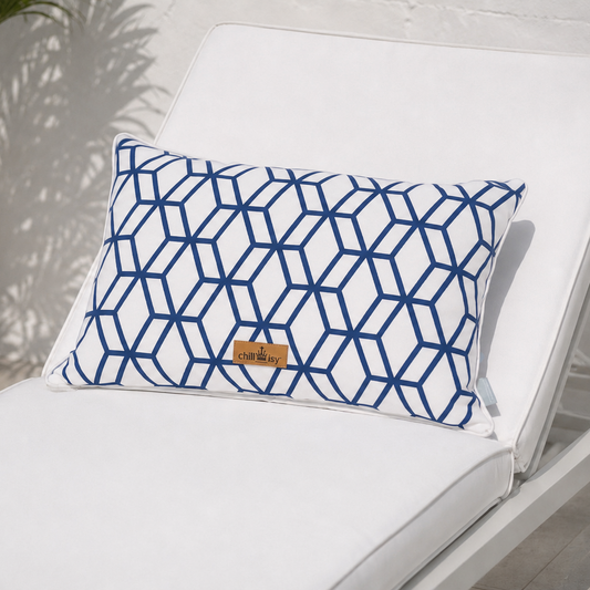 Luxuriöses Outdoor-Dekokissen mit weißem Grundstoff und marineblauem geometrischem Muster auf einer eleganten weißen Gartenlounge mit Meerblick. Das Kissen besitzt einen weißen Keder und das goldene chillisy®-Label und ist handgefertigt in Deutschland – ideal für Terrasse, Garten und Lounge.