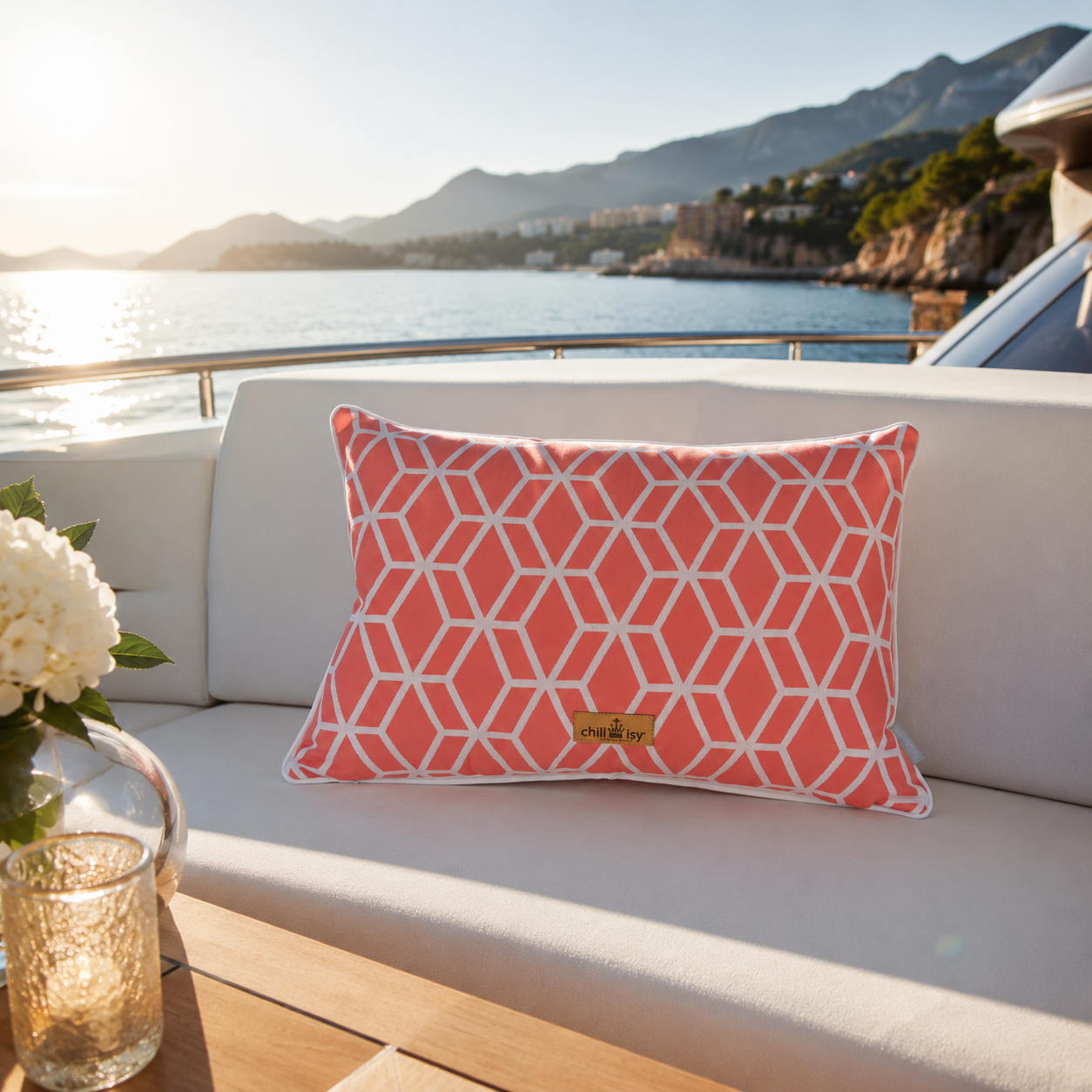 Luxuriöses chillisy® Outdoor-Kissen mit korallfarbenem geometrischem Muster, weißem Keder und goldenem chillisy®-Label auf einer eleganten weißen Yacht-Lounge mit Blick auf das Meer bei warmem Abendlicht.
