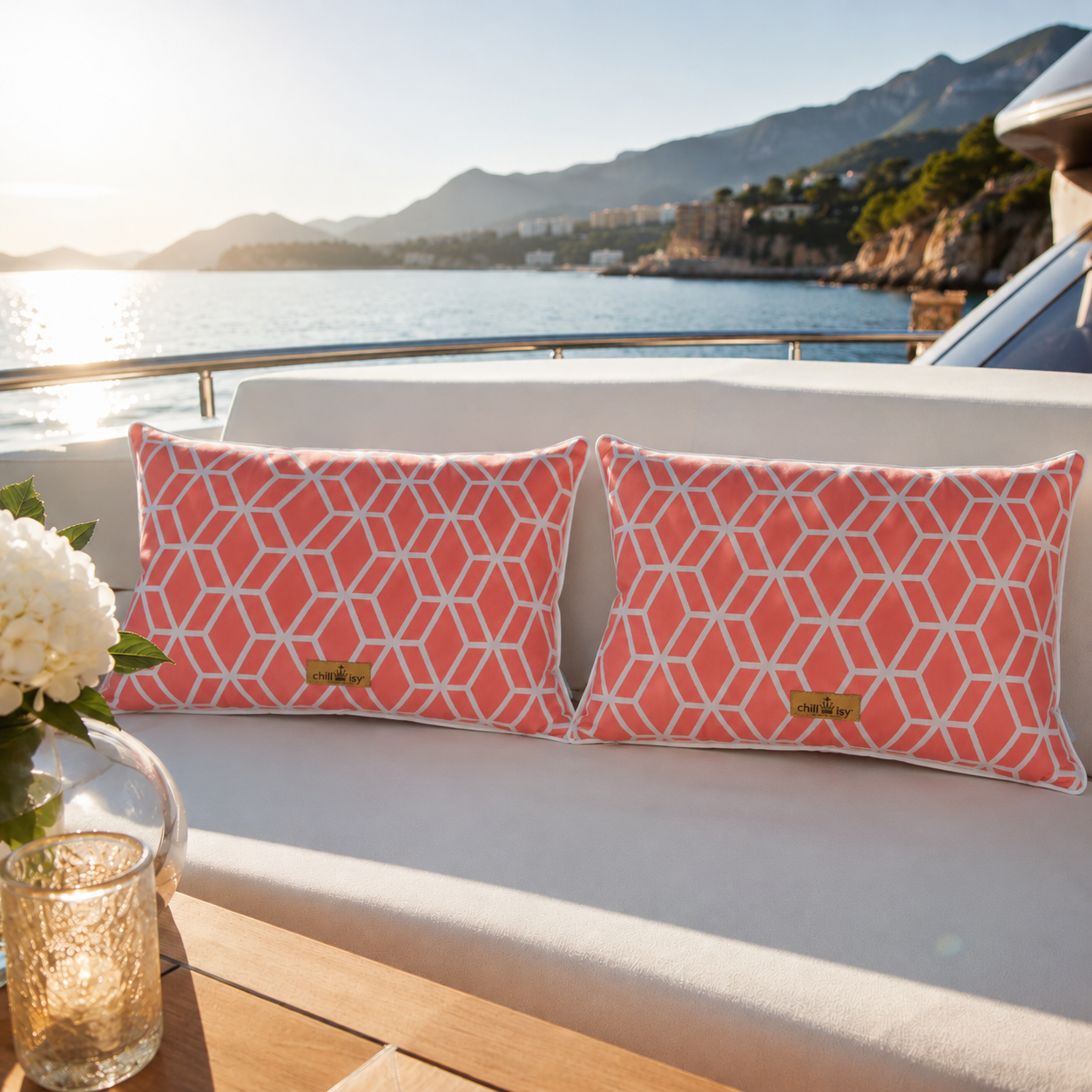 Zwei luxuriöse chillisy® Outdoor-Kissen mit korallfarbenem geometrischem Muster, weißem Keder und goldenem chillisy®-Label auf einer eleganten weißen Yacht-Lounge mit Blick auf das Meer bei warmem Abendlicht.