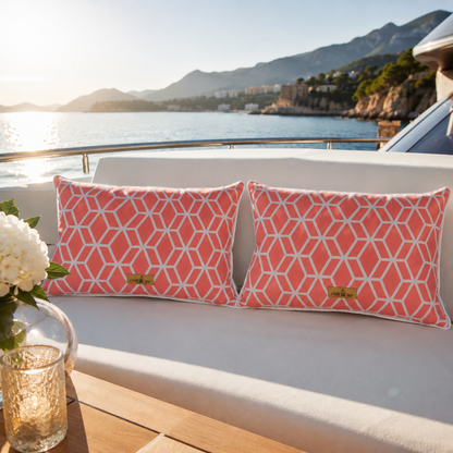 Zwei luxuriöse chillisy® Outdoor-Kissen mit korallfarbenem geometrischem Muster, weißem Keder und goldenem chillisy®-Label auf einer eleganten weißen Yacht-Lounge mit Blick auf das Meer bei warmem Abendlicht.
