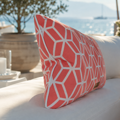 Detailaufnahme eines luxuriösen Outdoor-Dekokissens in Koralle mit weißem geometrischem Muster und weißem Keder auf einer eleganten weißen Gartenlounge. Im Hintergrund sorgen Meerblick und mediterranes Ambiente für eine exklusive Wohlfühlatmosphäre.
