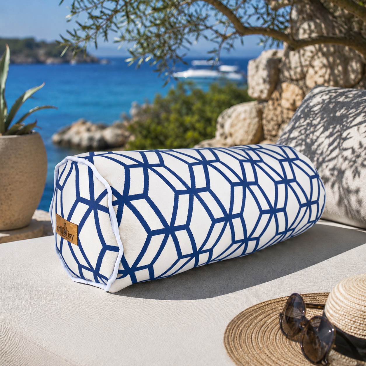 Outdoor-Nackenrolle von chillisy® mit weißem Stoff, marineblauem geometrischem Muster, weißem Keder und goldenem chillisy®-Label auf einer eleganten Gartenlounge am Meer. Luxuriöse Outdoor-Dekoration für Terrasse, Pool und Yacht mit mediterranem Küstenblick.
