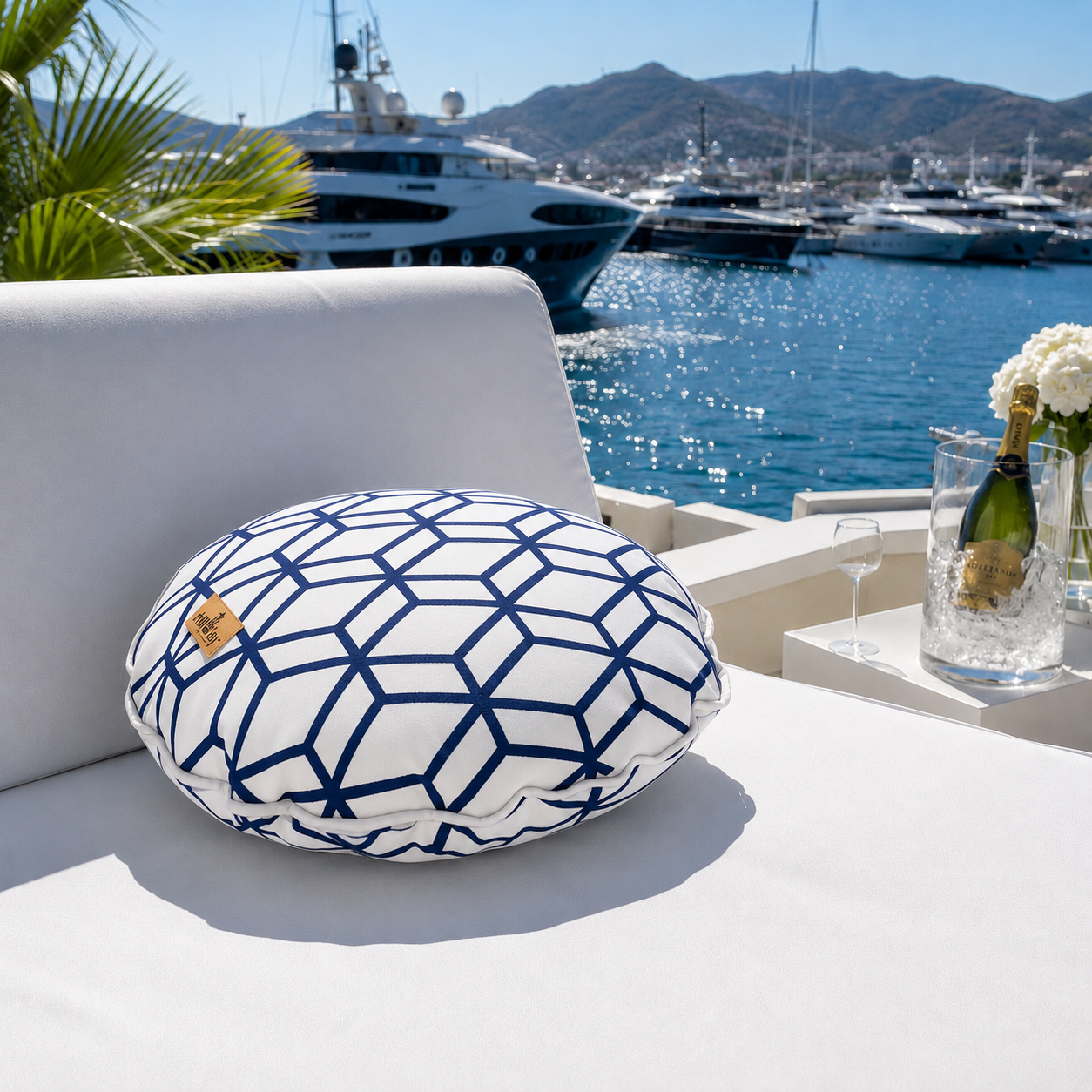 Rundes Outdoor-Dekokissen von chillisy® mit weißem Stoff, marineblauem geometrischem Muster, weißem Keder und goldenem chillisy®-Label auf einer eleganten weißen Gartenlounge im Yachthafen. Im Hintergrund glitzern luxuriöse Yachten, türkisblaues Wasser und ein Champagnerkühler – stilvolles Ambiente für Terrasse, Pool und Yacht.