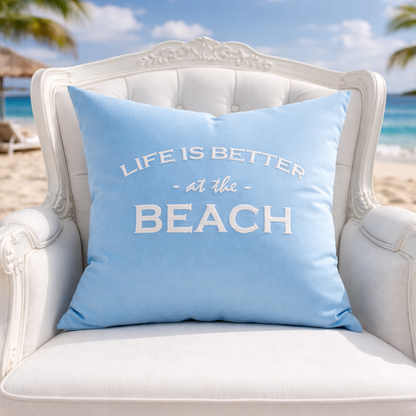 Dekokissen Aqua Blau mit weißer Stickerei LIFE IS BETTER AT THE BEACH auf weißem Sessel am Meer