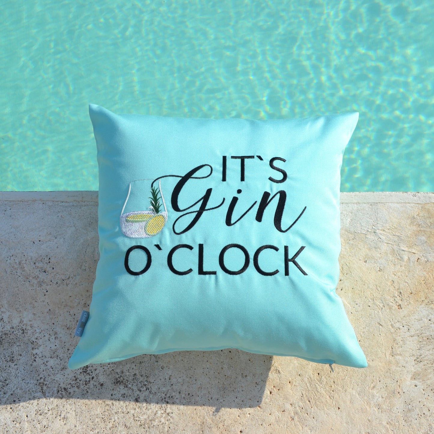 Mint farbenes Kissen bestickt mit dem Motiv IT‘S GIN O‘CLOCK abgebildet auf Terrasse am Wasser.