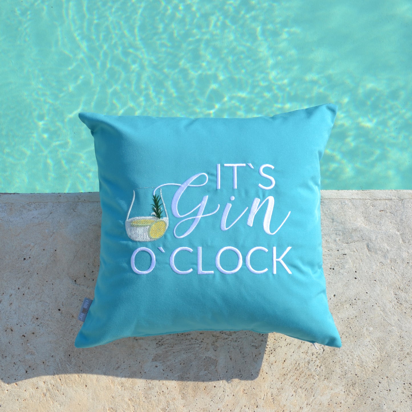 Türkises Kissen mit weißem Stick IT'S GIN O'CLOCK und Gin Tonic Glas auf Terrassenboden am Pool liegend.