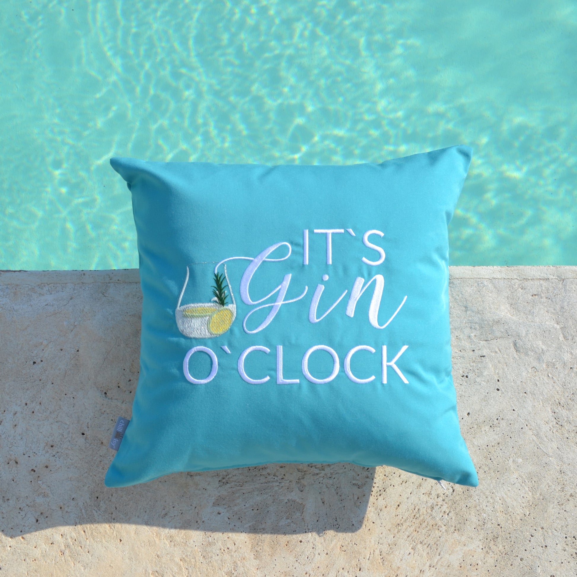 Türkises Kissen mit weißem Stick IT'S GIN O'CLOCK und Gin Tonic Glas auf Terrassenboden am Pool liegend.
