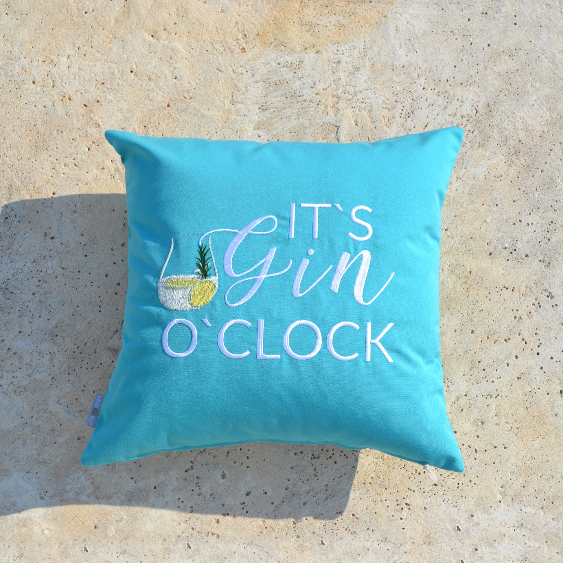 Türkises Kissen mit weißem Stick IT'S GIN O'CLOCK und Gin Tonic Glas auf Terrassenboden liegend.