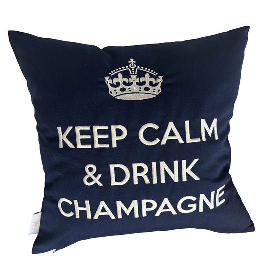 Dunkelblaues Dekokissen mit weißer Stickerei einer Krone und dem Schriftzug „KEEP CALM & DRINK CHAMPAGNE“, liegend auf einem hellen Ledersessel.