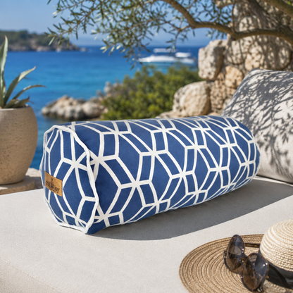 chillisy® Outdoor-Nackenrolle mit blau-weißem geometrischem Würfelmuster und weißer Kederumrandung auf einer luxuriösen Sonnenliege am Meer. Mediterrane Natur, türkisblaues Wasser und Natursteinmauern schaffen ein exklusives Ambiente – hochwertiges Designkissen für Garten, Terrasse und Outdoor-Lounge.