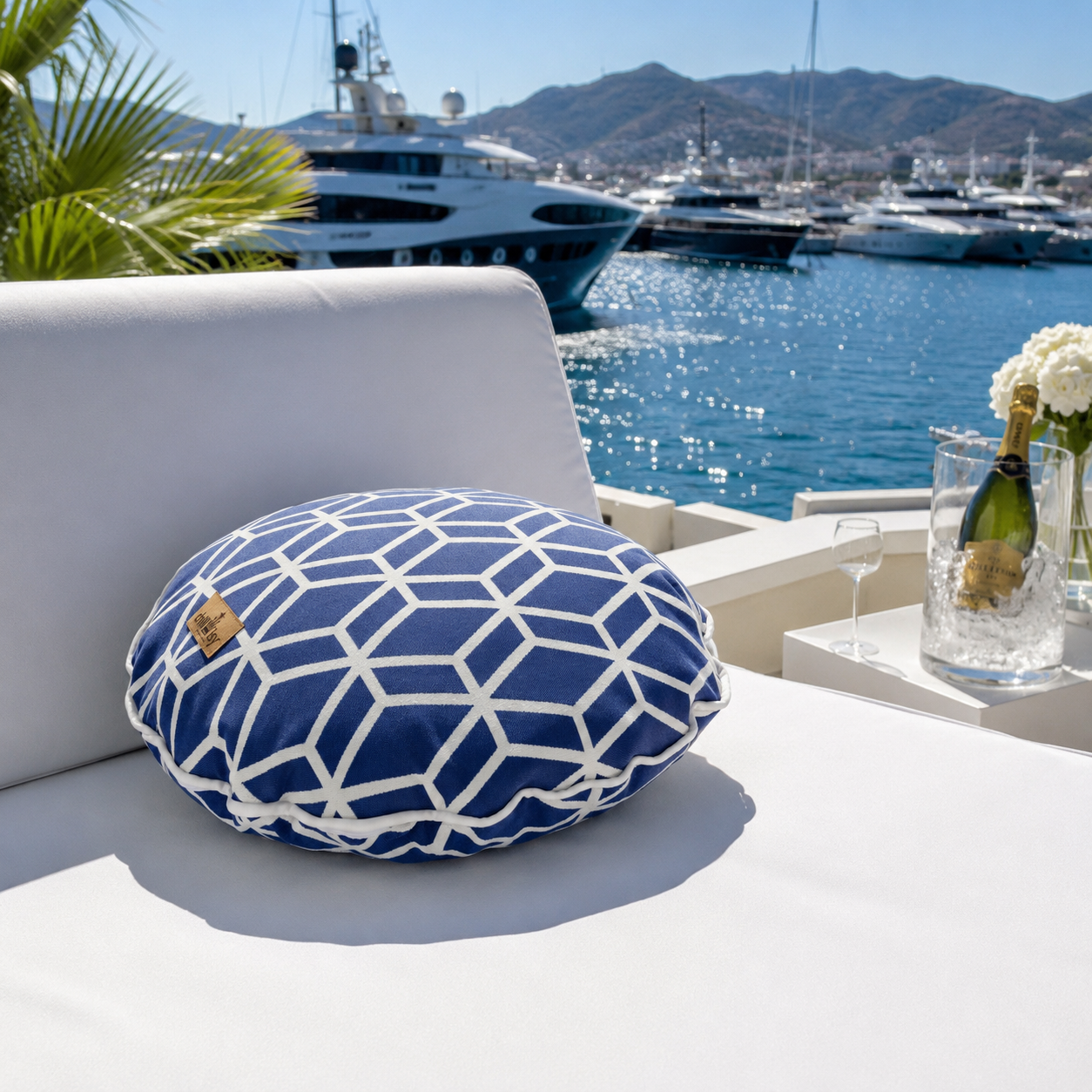 Rundes chillisy® Outdoor-Dekokissen mit blau-weißem geometrischem Würfelmuster und weißer Kederumrandung auf einer eleganten weißen Gartenlounge. Im Hintergrund eine luxuriöse Yacht mit türkisblauem Meer und Sonnenschein – exklusives Designerkissen für Terrasse, Balkon und Garten.