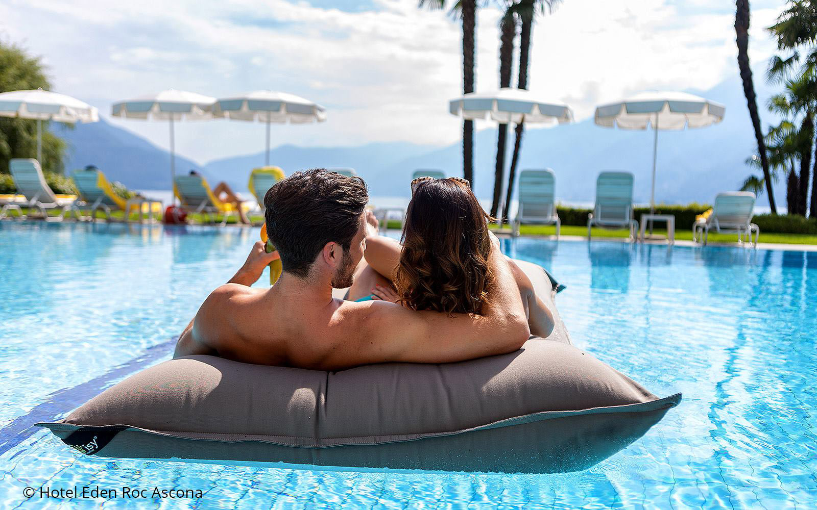 Premium Poolkissen Maxi 190x130 cm von chillisy in der Farbe Taupe im Hotelpool mit zwei Erwachsenen darauf liegend.