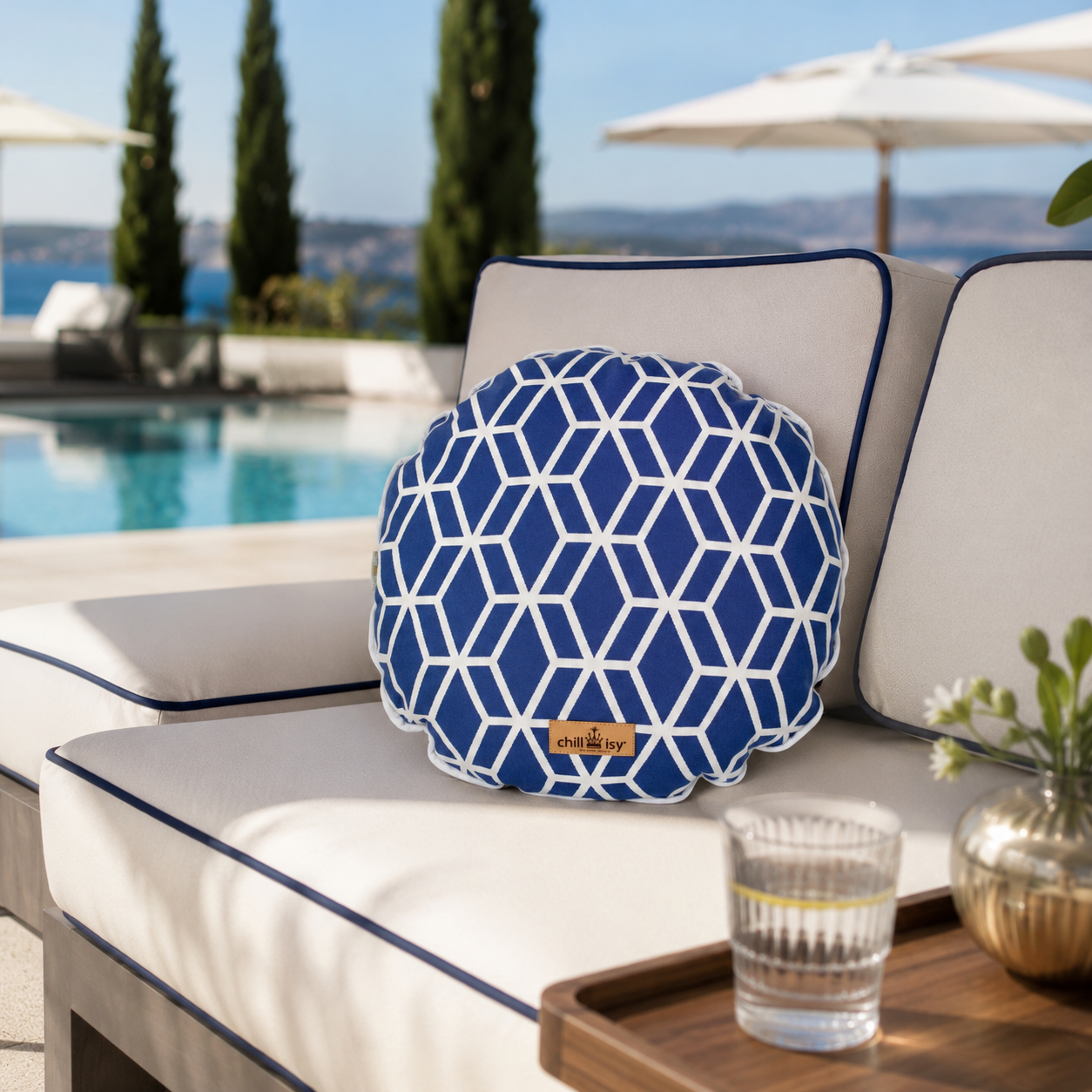 Rundes chillisy® Outdoor-Dekokissen mit blau-weißem geometrischem Würfelmuster und weißer Kederumrandung auf einer eleganten weißen Gartenlounge am Pool. Im Hintergrund sorgen ein türkisblauer Swimmingpool, mediterrane Zypressen und Sonnenschirme für luxuriöses Resort-Ambiente – hochwertiges Designerkissen für Garten, Terrasse und Outdoor-Lounge.